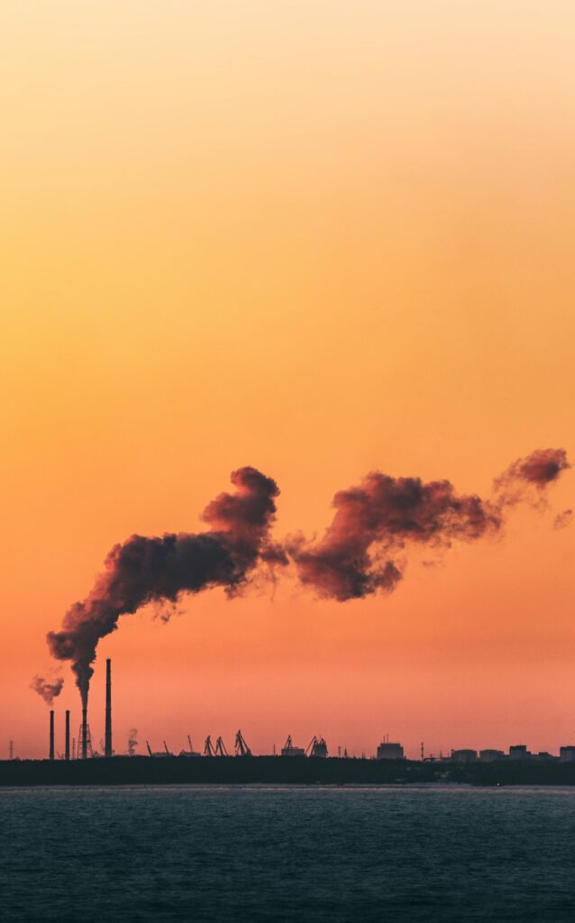 Orange sky at sunset with industrial smokestacks emitting smoke over water.